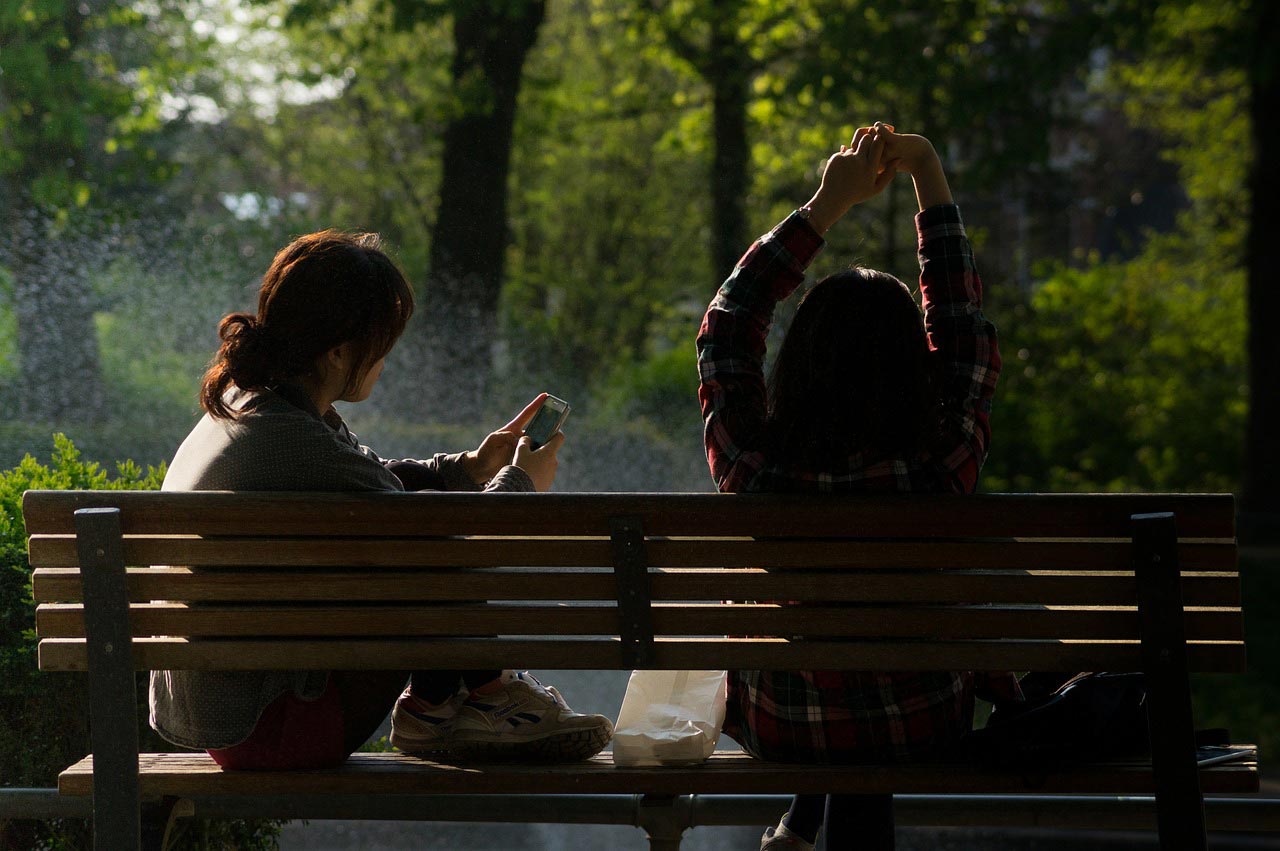 Two women sit on a park bench at the park on a summer day.