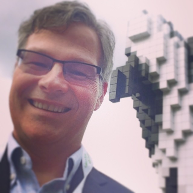 A man is on the left, and we see him from the neck up. He has glasses with black frames, grey hair, and is smiling widely. Behind him there is a sculpture of an orca in breach position, which is out of focus, and seems to be made up of black and white squares like lego.