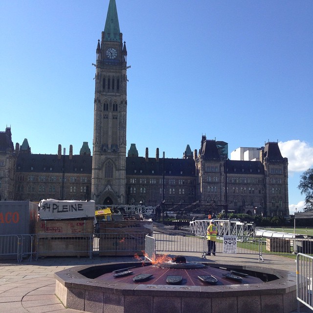 A view of Centre Block, Peace Tower -- a low colonial style building made of stone with a tall clock tower slightly left of center --, with Centennial Flame in foreground. The Centennial Flame is a low cylindrical black concrete and metal structure with a torch in the middle.