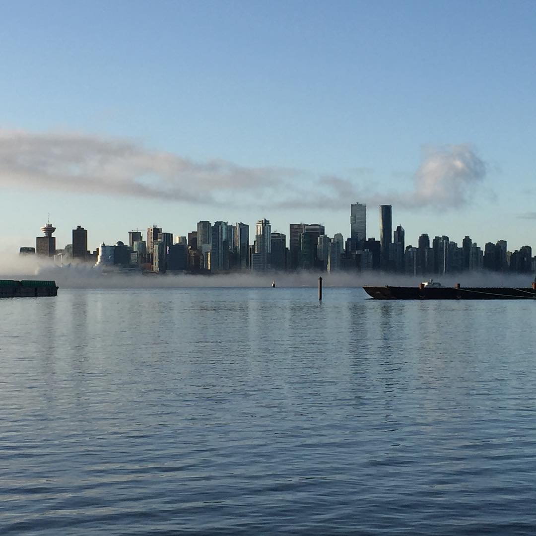 Looking across the calm ocean to downtown vancouver. The sky is blue and there is a low fog cloud along the line where downtown meets to ocean on the otherside.