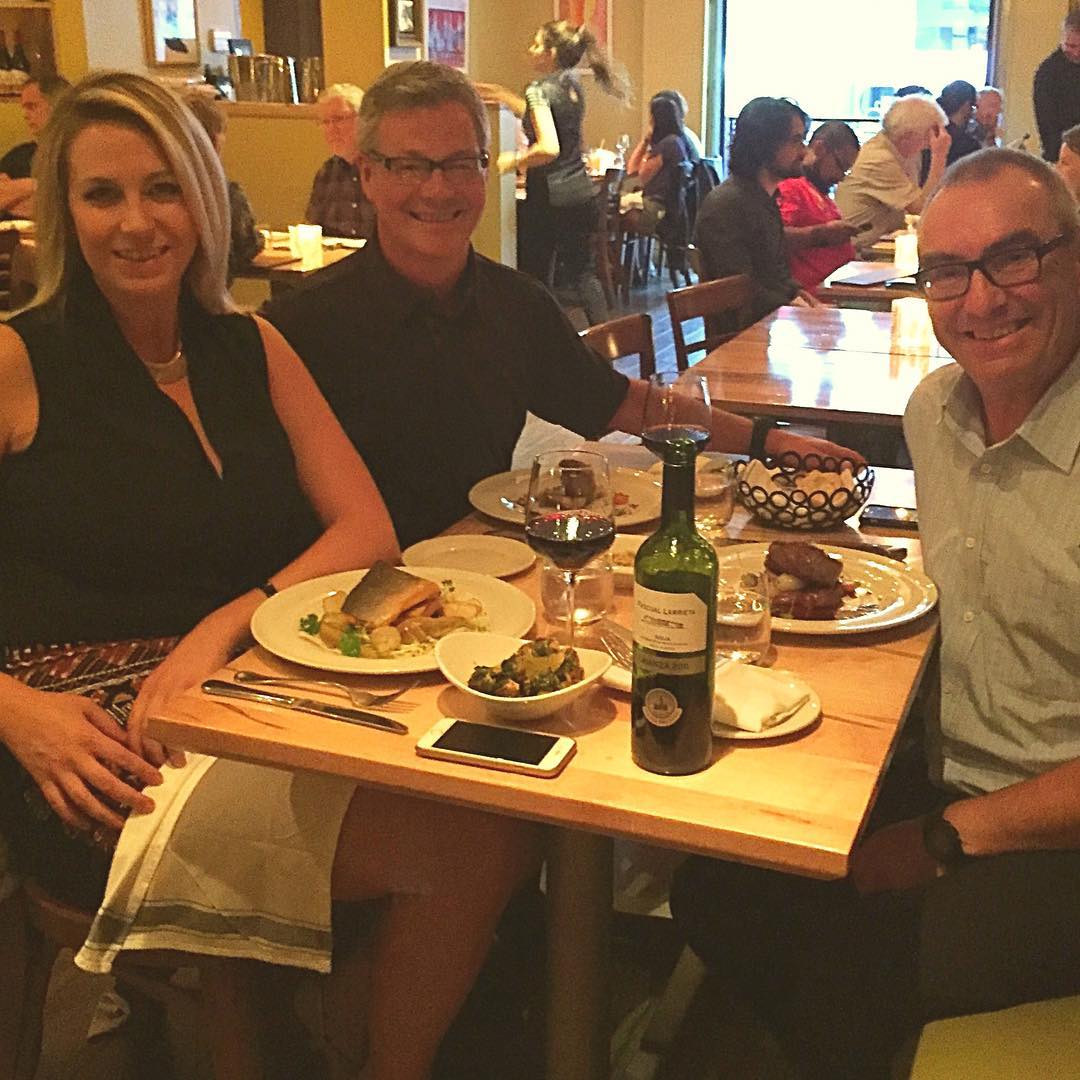 Three people sitting around a square, light brown wooden table covered with food. On the right side is a man with grey hair and black glasses who is smiling. On the other side are a woman with shoulder length blonde hair, and a man with grey hair and black glasses who are also both smiling.