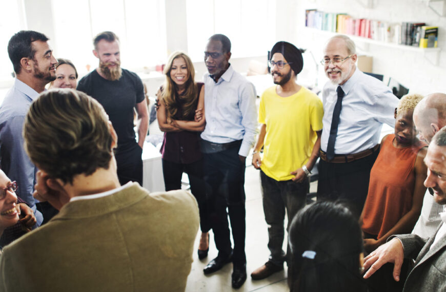 A group of people stand in a circle in an office having a meeting and smiling and laughing together.