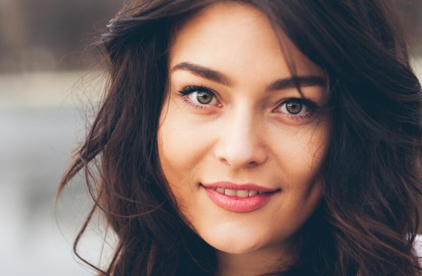 A young woman smiles and slightly tilts her head, standing outside in front of a lake.