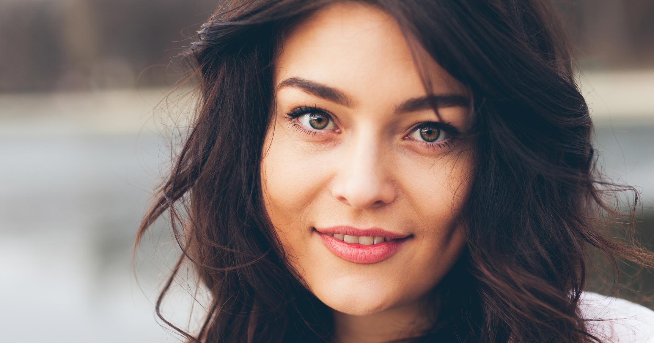 A young woman smiles and slightly tilts her head, standing outside in front of a lake.
