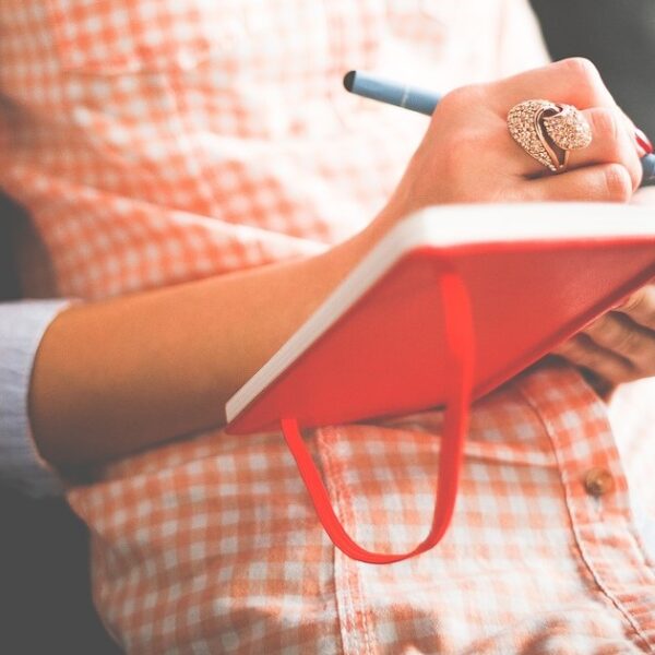 A close up of a person wearing an orange checkered button up top and writing in an orange notebook. It is only a photo of their torso and arms.