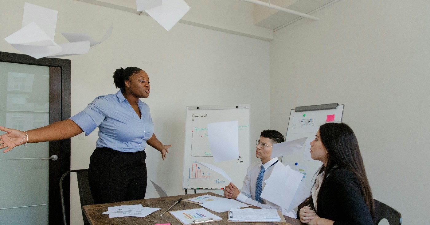 A Black woman throws papers in the air while her two white coworkers look on from their conference room table.